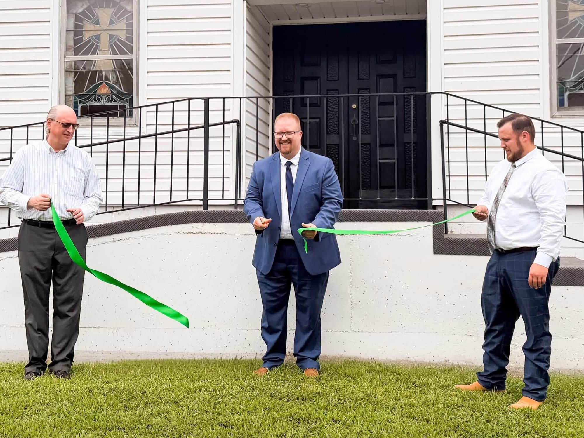 Three men holding a green ribbon across the front steps of the church on charter Sunday, moments before it was cut