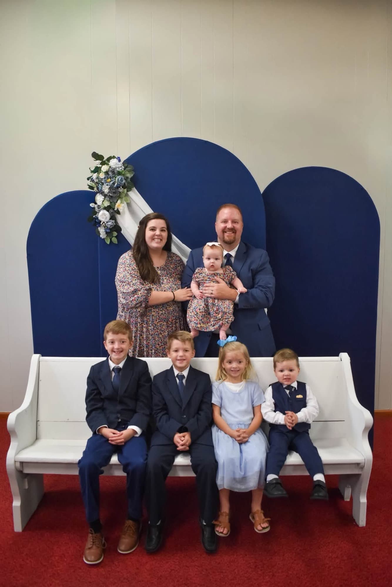 Pastor Nick Brown and his wife Sam standing behind a white pew with their five children, dressed for church in front of navy backdrops