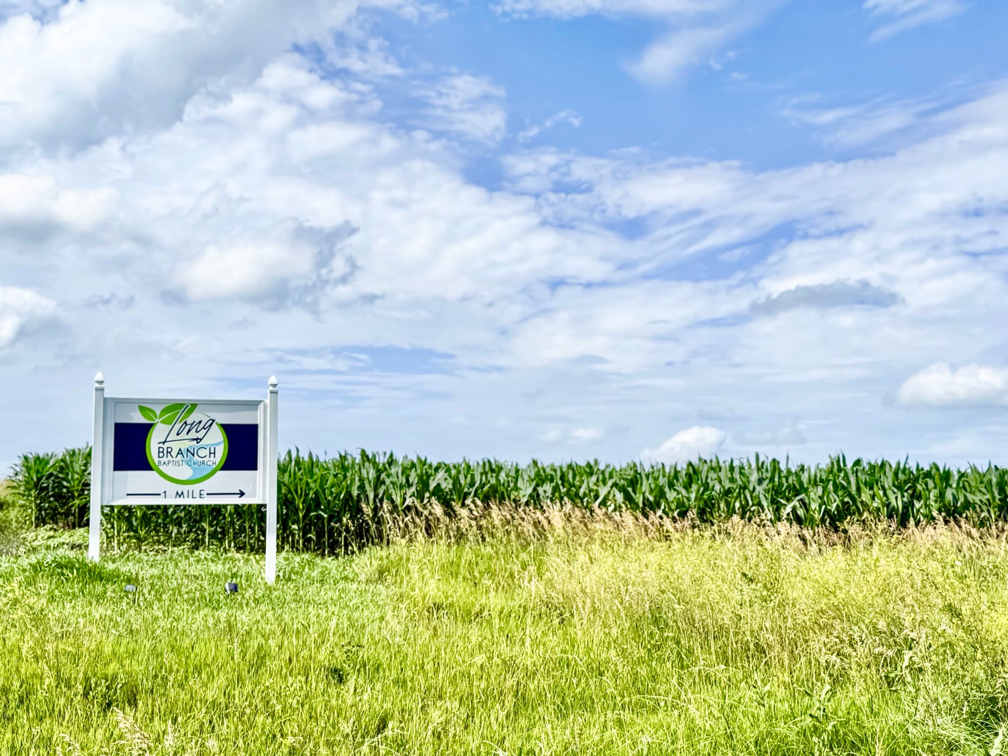 The church's road sign at the edge of a cornfield, with an arrow reading one mile